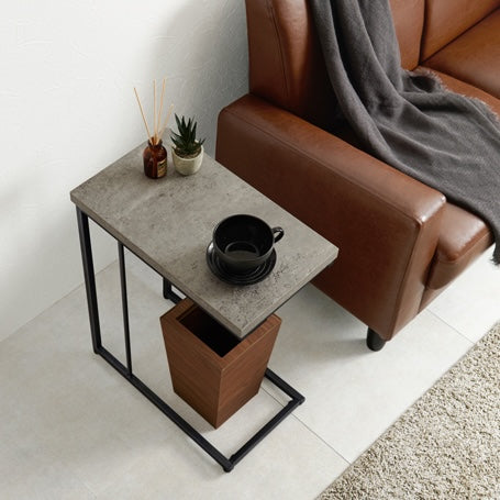 リビングでブラウンの革製ソファの隣に置いてあるコンクリ風デザインのサイドテーブルの下にゴミ箱が設置されている部屋の様子