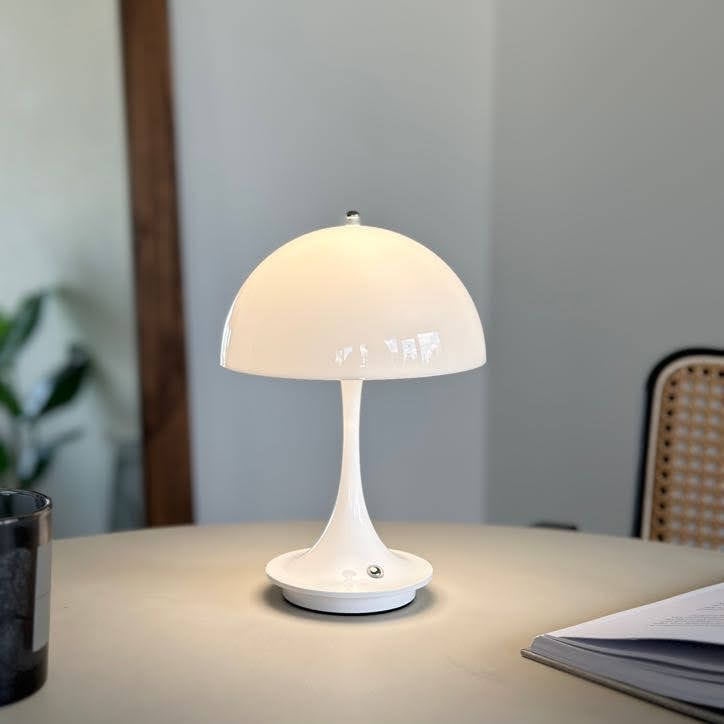 テーブルの上に置いたホワイトの大きめのシェードにくびれがかった支柱のデザインのランプが点灯している様子