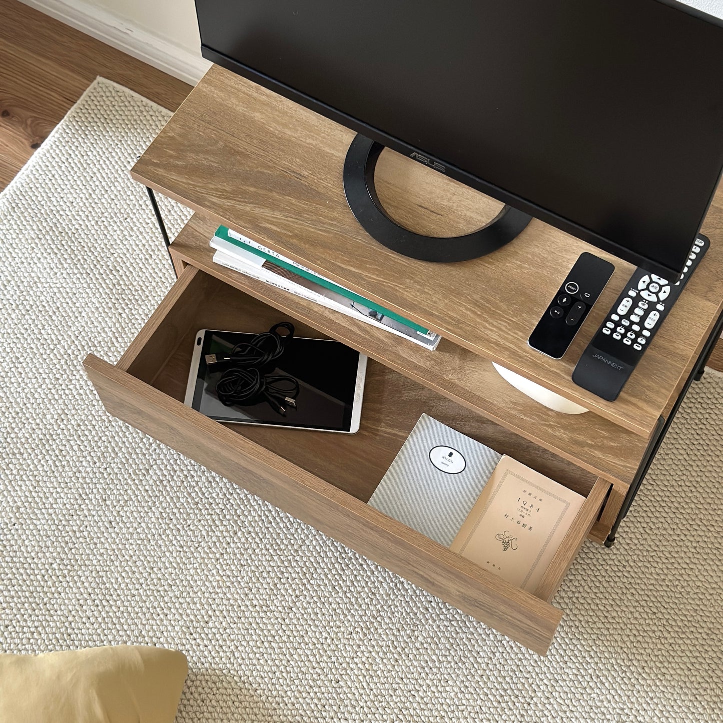 木目のウッドと細いブラックフレームのテレビボードの引き出し収納部分にタブレットとコード、文庫本が収納されている様子
