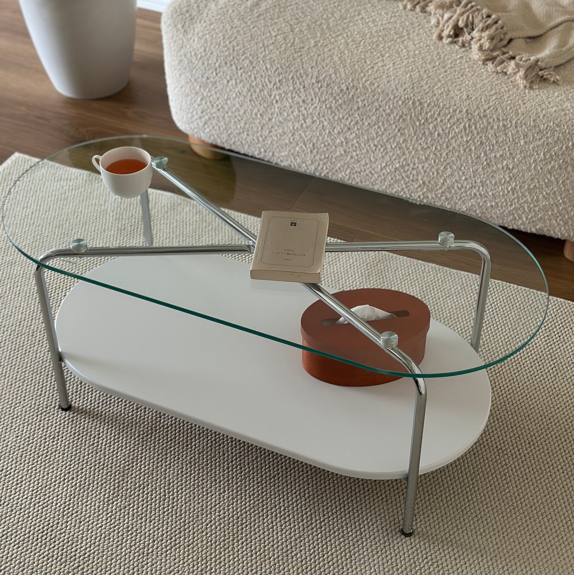 丸みのあるガラス天板のローテーブルの天板上にはコップと文庫本が置かれており、下の棚にはティッシュケースが置かれてある