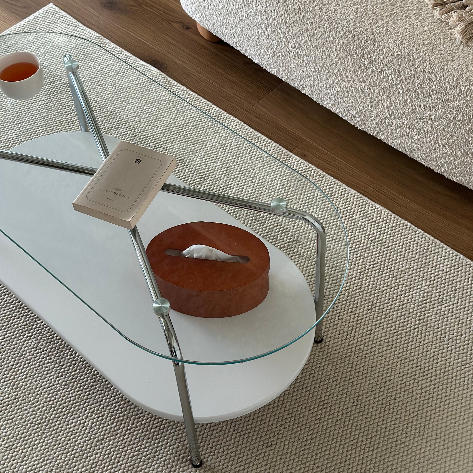 丸みのあるガラス天板のローテーブルの天板上にはコップと文庫本が置かれており、下の棚にはティッシュケースが置かれてある