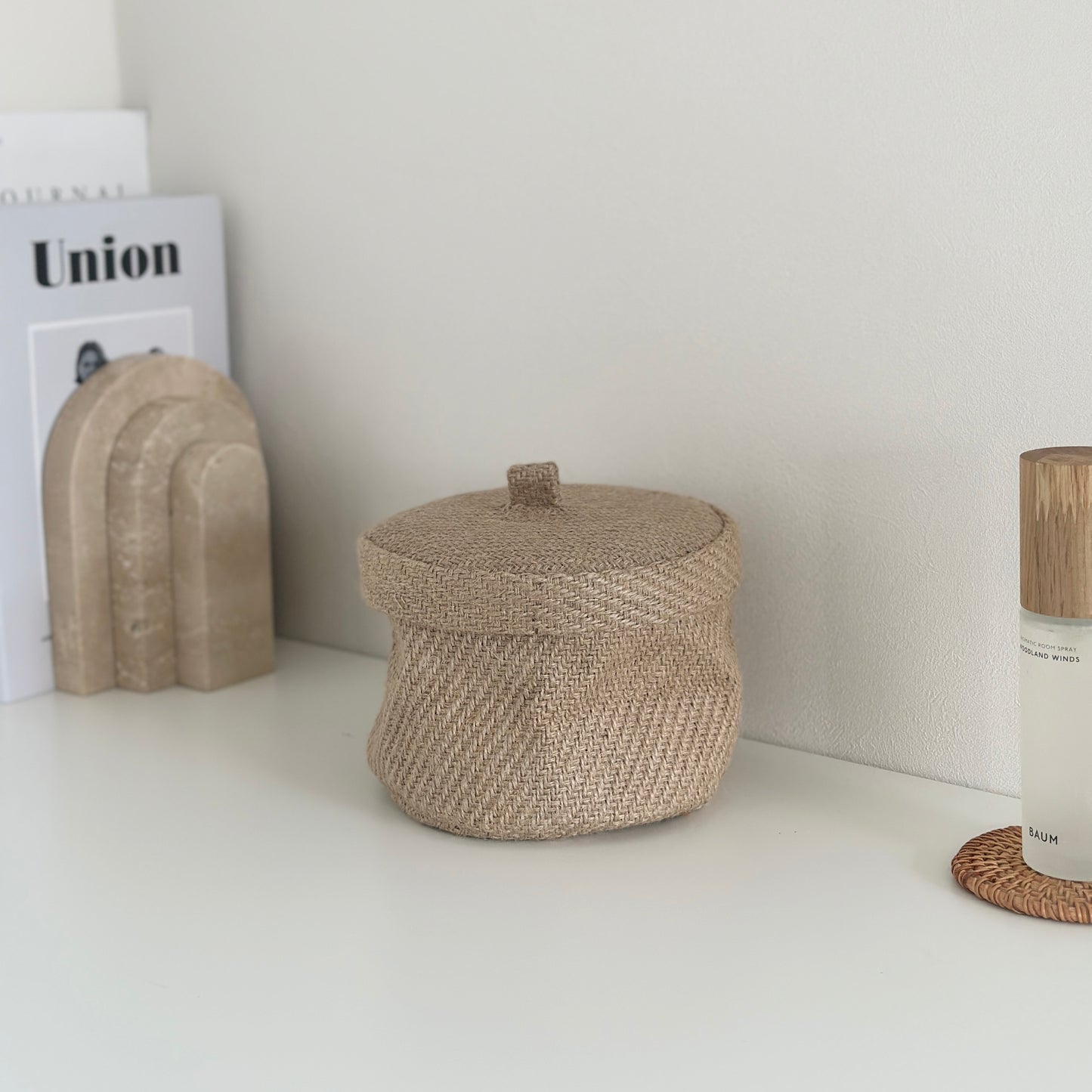 蓋が閉まった状態のリネン素材で出来た蓋付きのバスケットとその近くに化粧品や本が置かれている