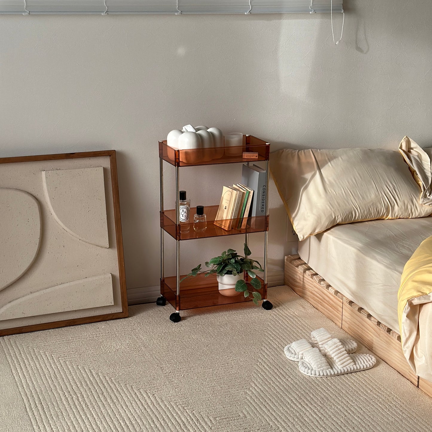 クリアなブラウンの3段シェルフにティッシュケースやキャンドル、香水、本、植物が収納され、ベッドサイドに置かれてある様子