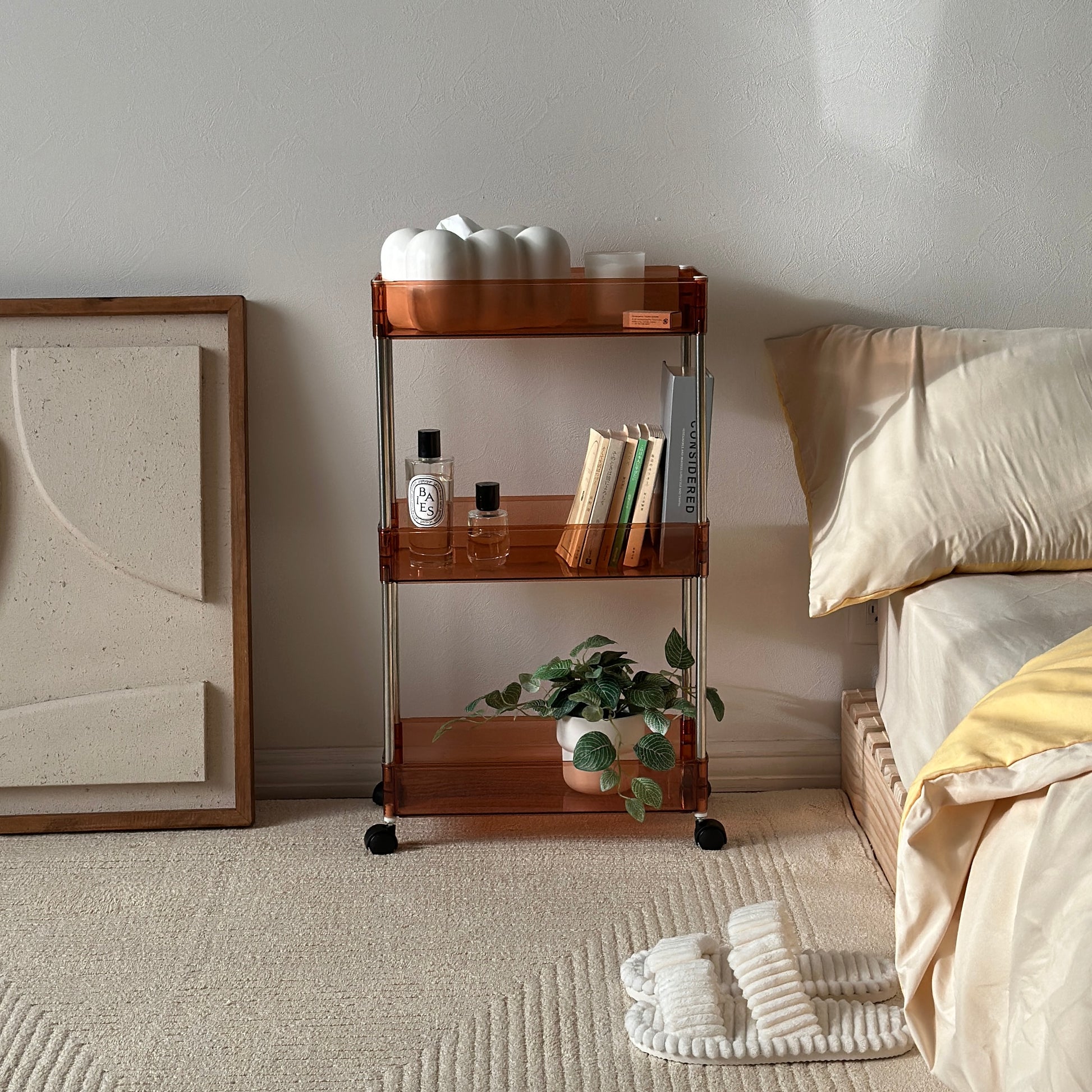 クリアなブラウンの3段シェルフにティッシュケースやキャンドル、香水、本、植物が収納され、ベッドサイドに置かれてある様子