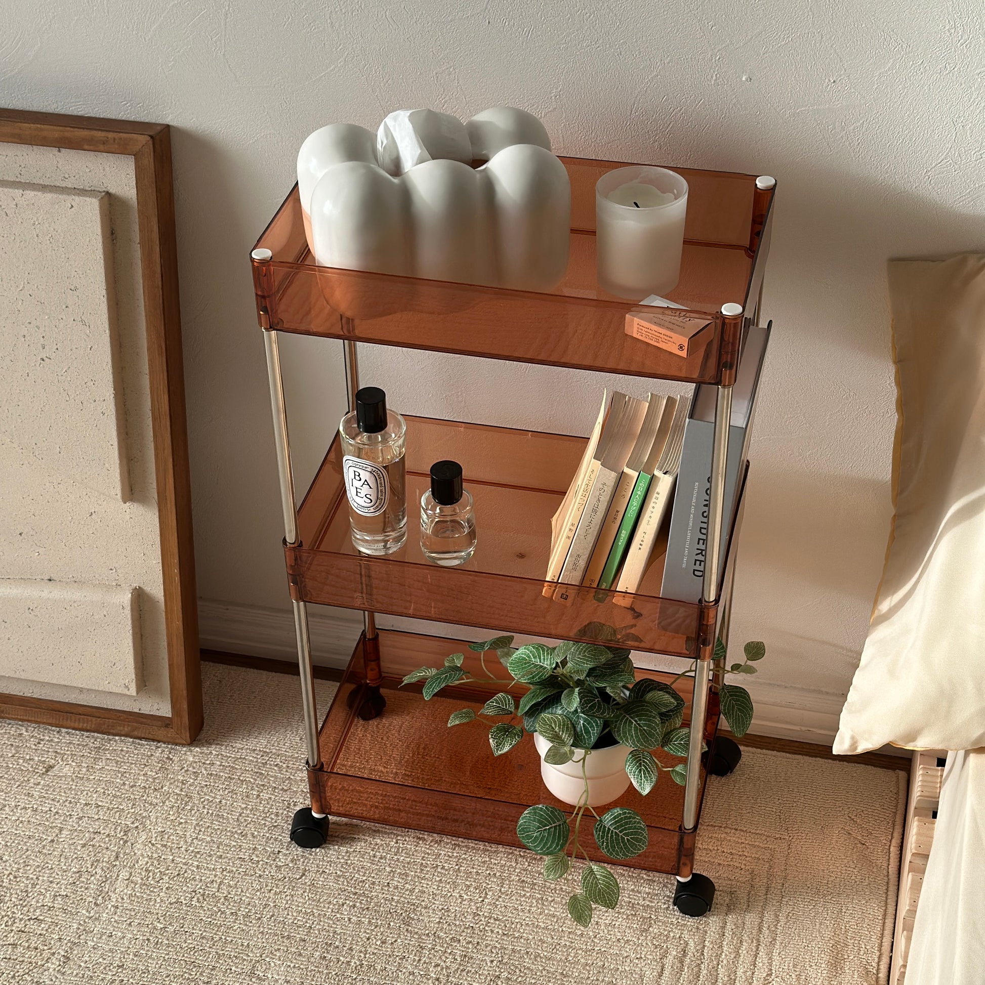 クリアなブラウンの3段シェルフにティッシュケースやキャンドル、香水、本、植物が置かれてある様子