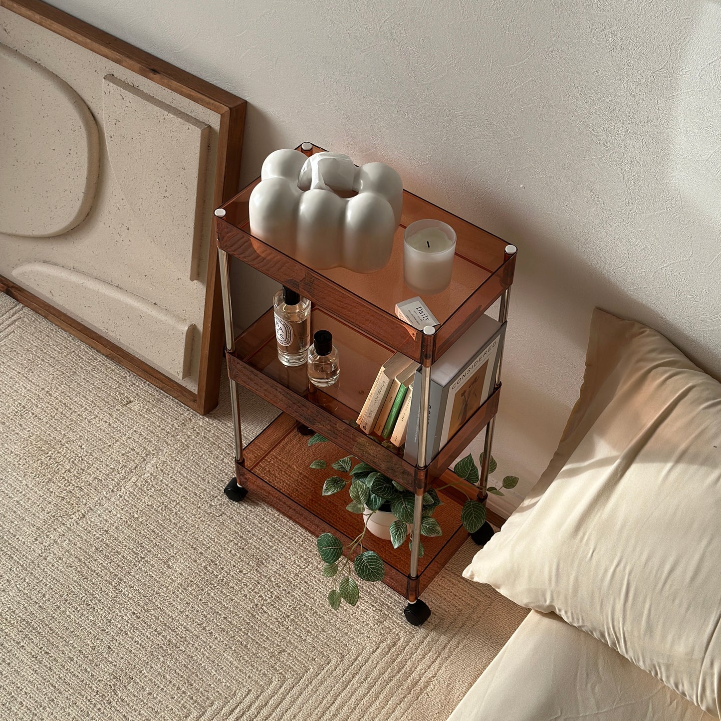 クリアなブラウンの3段シェルフにティッシュケースやキャンドル、香水、本、植物が置かれてある様子