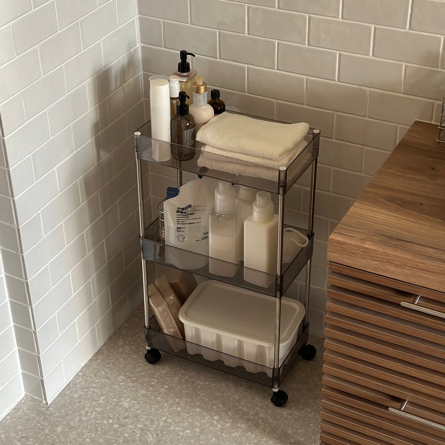 クリアなブラックの3段シェルフにスキンケア用品やタオル、洗濯洗剤などが収納され、洗面所に置かれてある様子