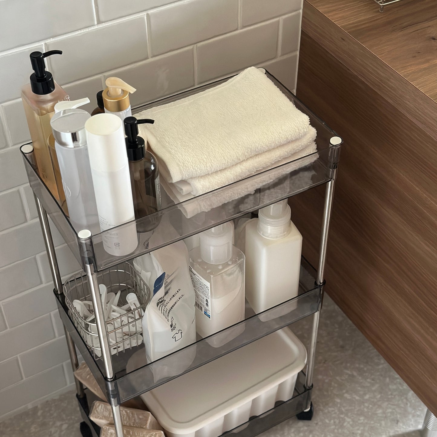 クリアなブラックの3段シェルフにスキンケア用品やタオル、洗濯洗剤などが収納され、洗面所に置かれてある様子