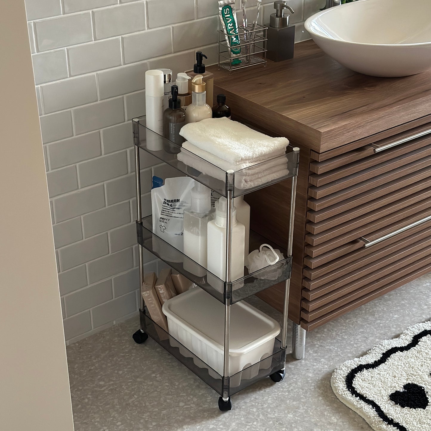 クリアなブラックの3段シェルフにスキンケア用品やタオル、洗濯洗剤などが収納され、洗面所に置かれてある様子