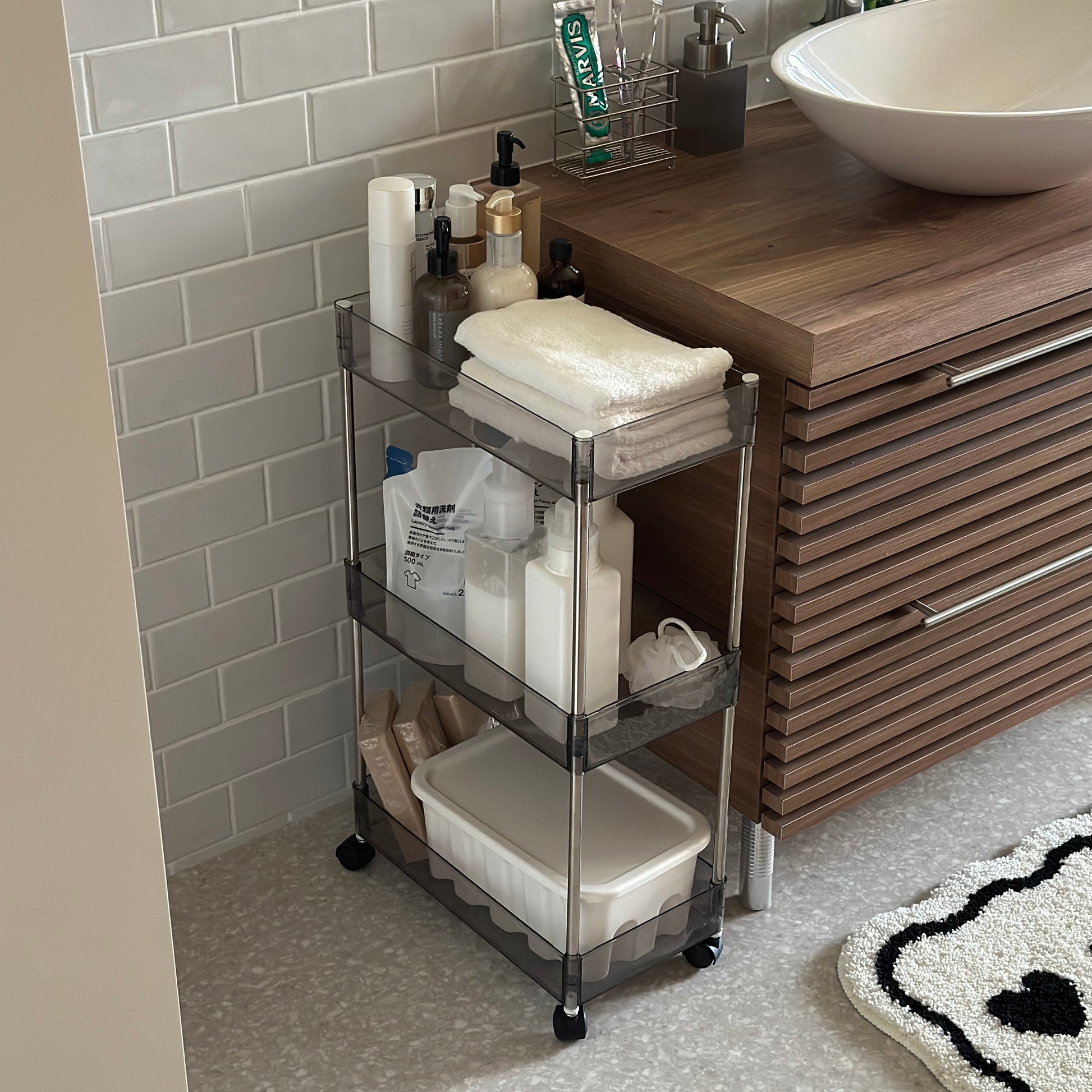 クリアなブラックの3段シェルフにスキンケア用品やタオル、洗濯洗剤などが収納され、洗面所に置かれてある様子