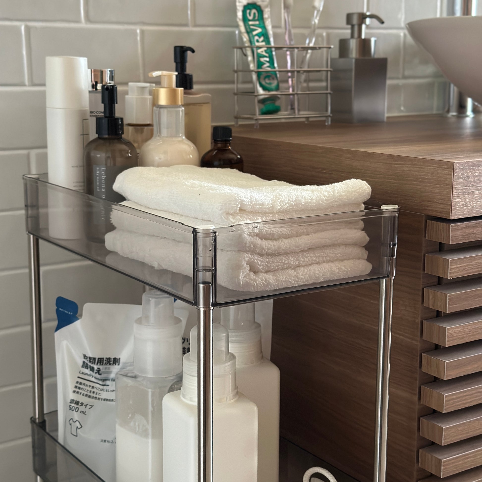 クリアなブラックの3段シェルフにスキンケア用品やタオル、洗濯洗剤などが収納され、洗面所に置かれてある様子