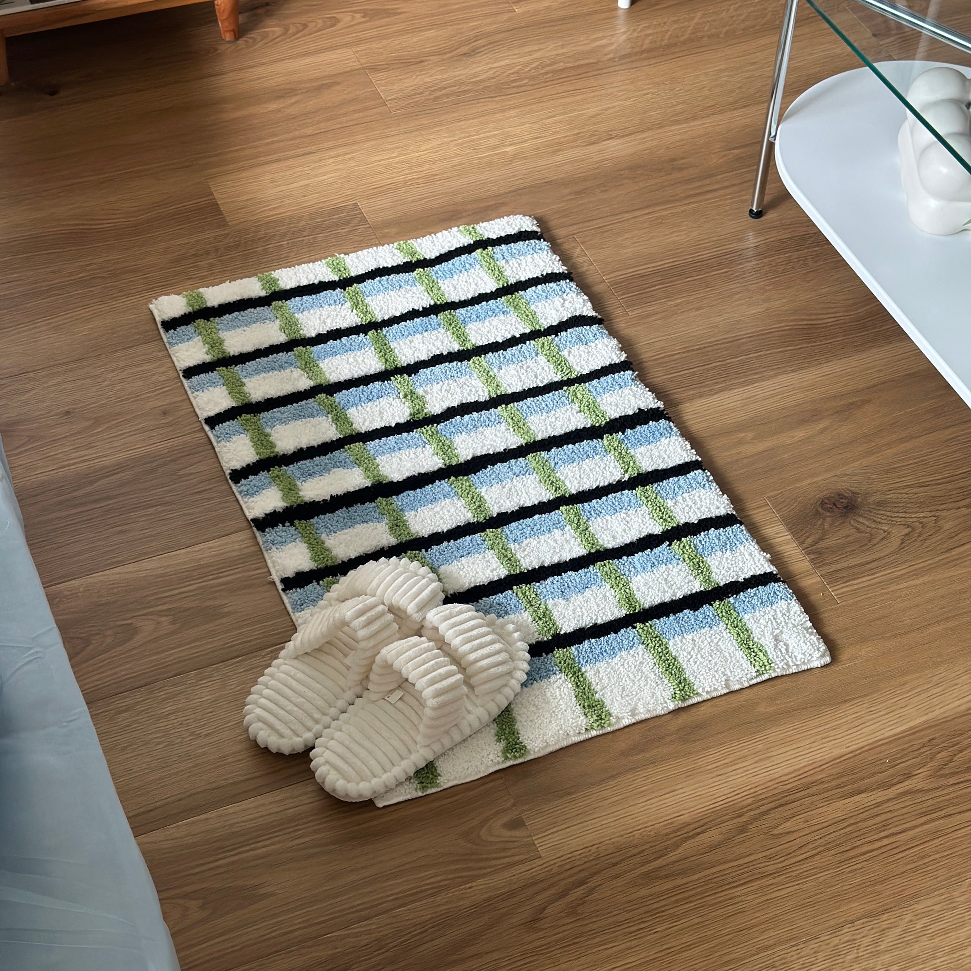 ブラックとグリーンとブルーの3色が入ったチェック柄のミニラグの上にホワイトのスリッパが置かれている様子