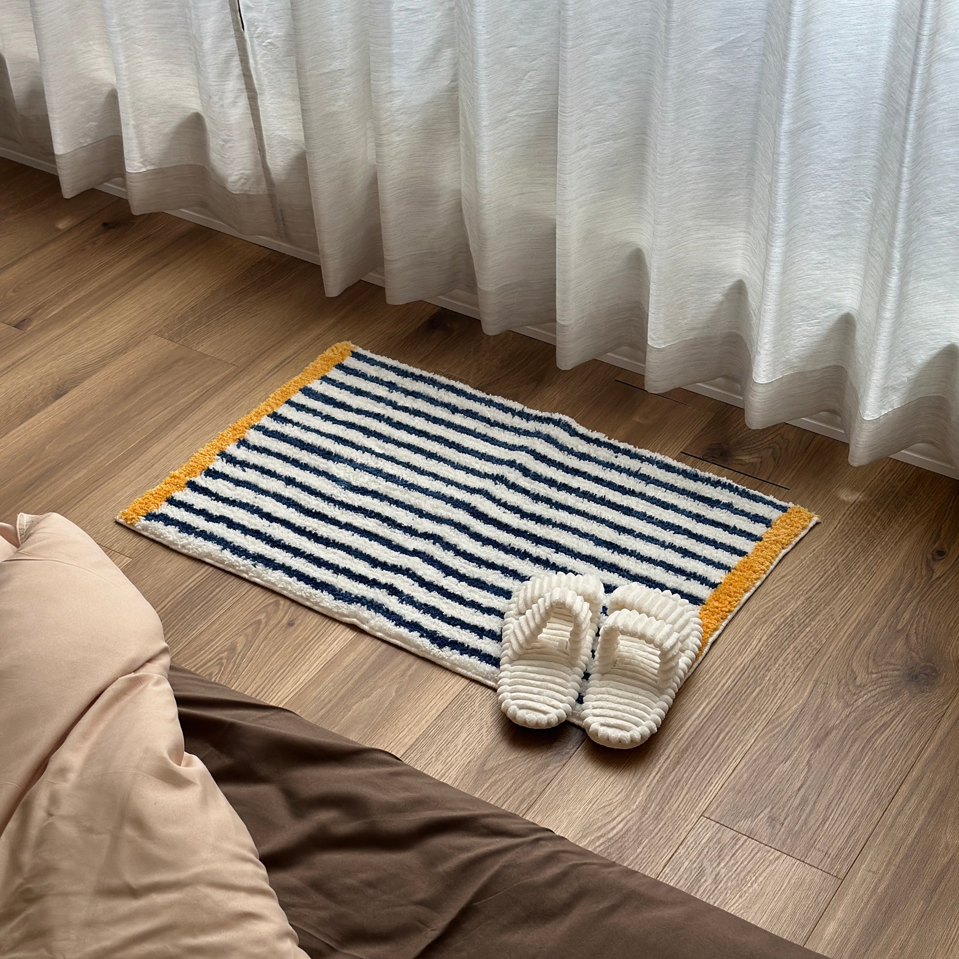 ブルーのボーダーとイエローの縁取りのミニラグの上にホワイトのスリッパが置かれている様子