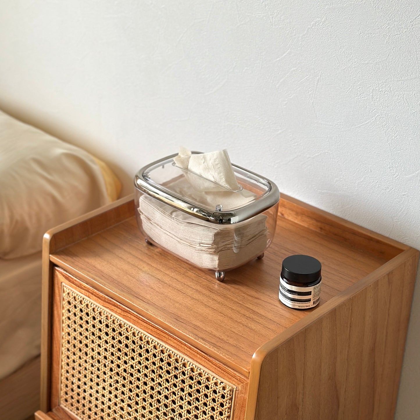 茶色い木目の棚の上にシルバーのラインが入った透明な脚付ティッシュケースが置かれてある。