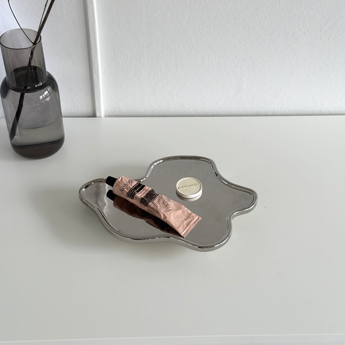 縁がウェーブ状になっている脚付きのシルバープレートの上にハンドクリームが乗っていて後ろに植物が写っている