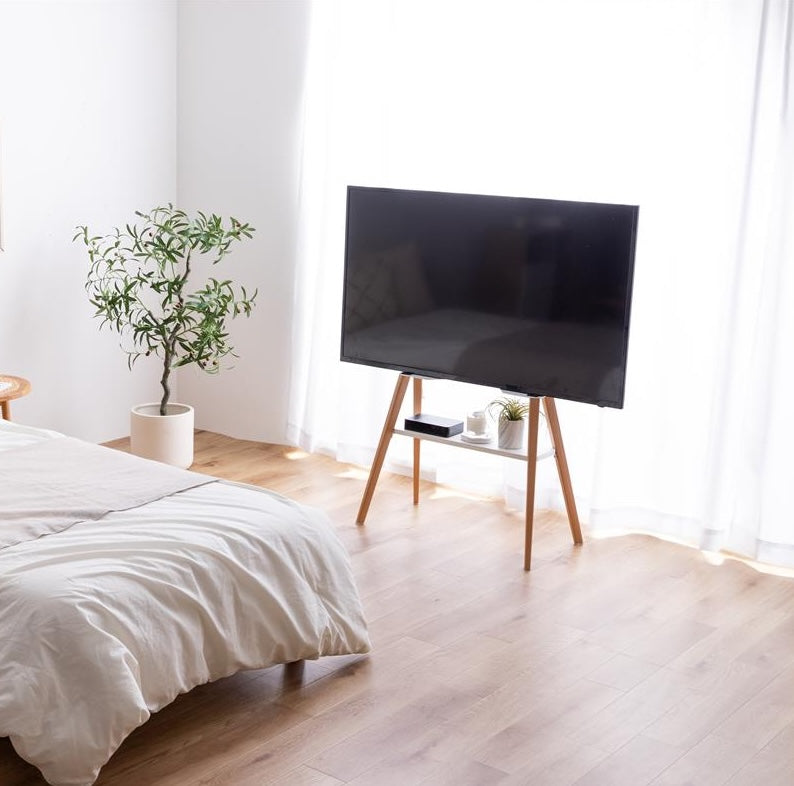 ホワイトのラックが付いたウッド脚のテレビスタンドがベッド横に置かれている様子
