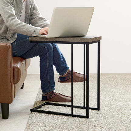 リビングでブラウンの革製ソファに座ってコンクリ風デザインのサイドテーブルの上でパソコン作業をしている人の様子