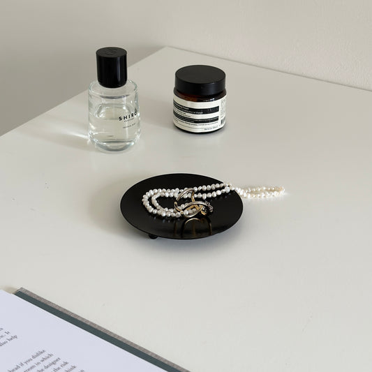 脚付のブラックの鉄製の丸いプレートの上にアクセサリーが置かれていて周りに香水や化粧品が置かれている