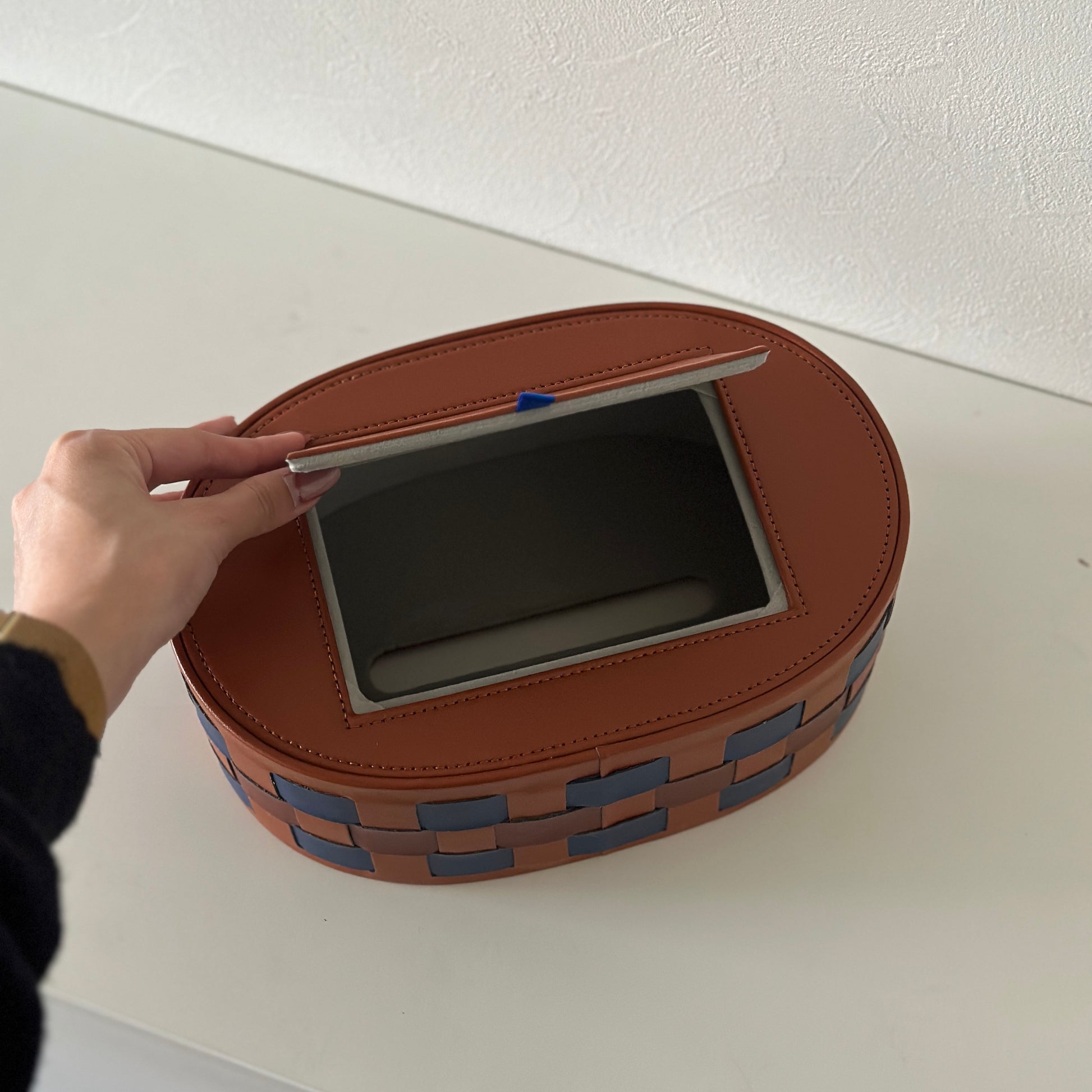 ブルーとブラウンの編み込みデザインのオーバル型のティッシュケースの蓋を開けている様子