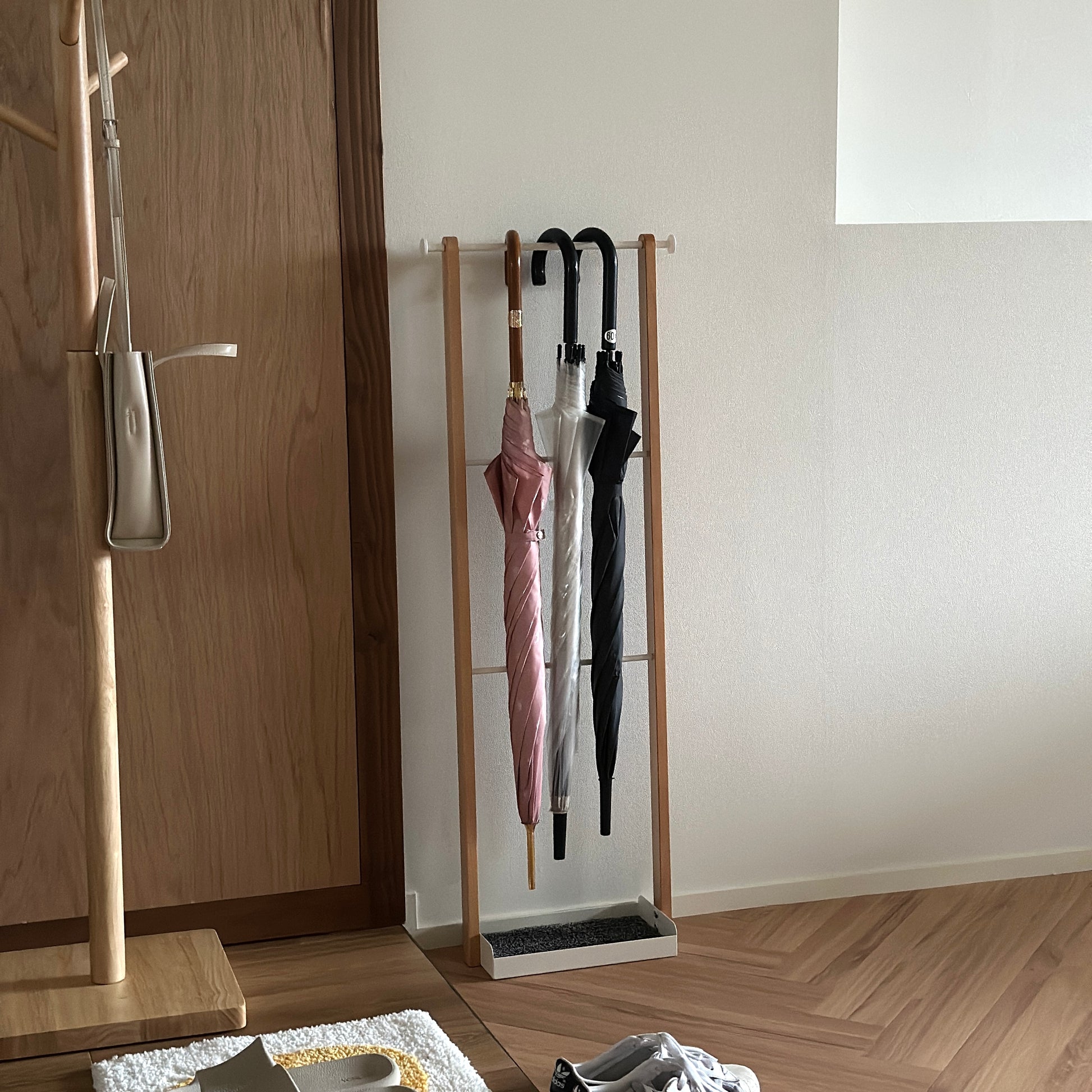 部屋の中に天然木とホワイトスチールの3段のアンブレララックが置かれており、ピンクの傘とビニール傘、黒の傘がかけられている。