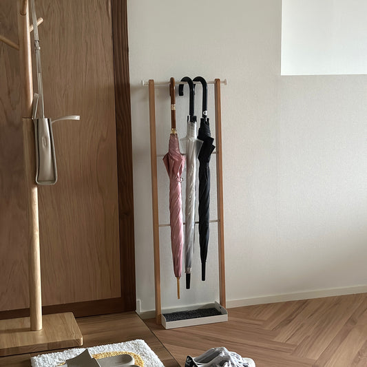 部屋の中に天然木とホワイトスチールの3段のアンブレララックが置かれており、ピンクの傘とビニール傘、黒の傘がかけられている。