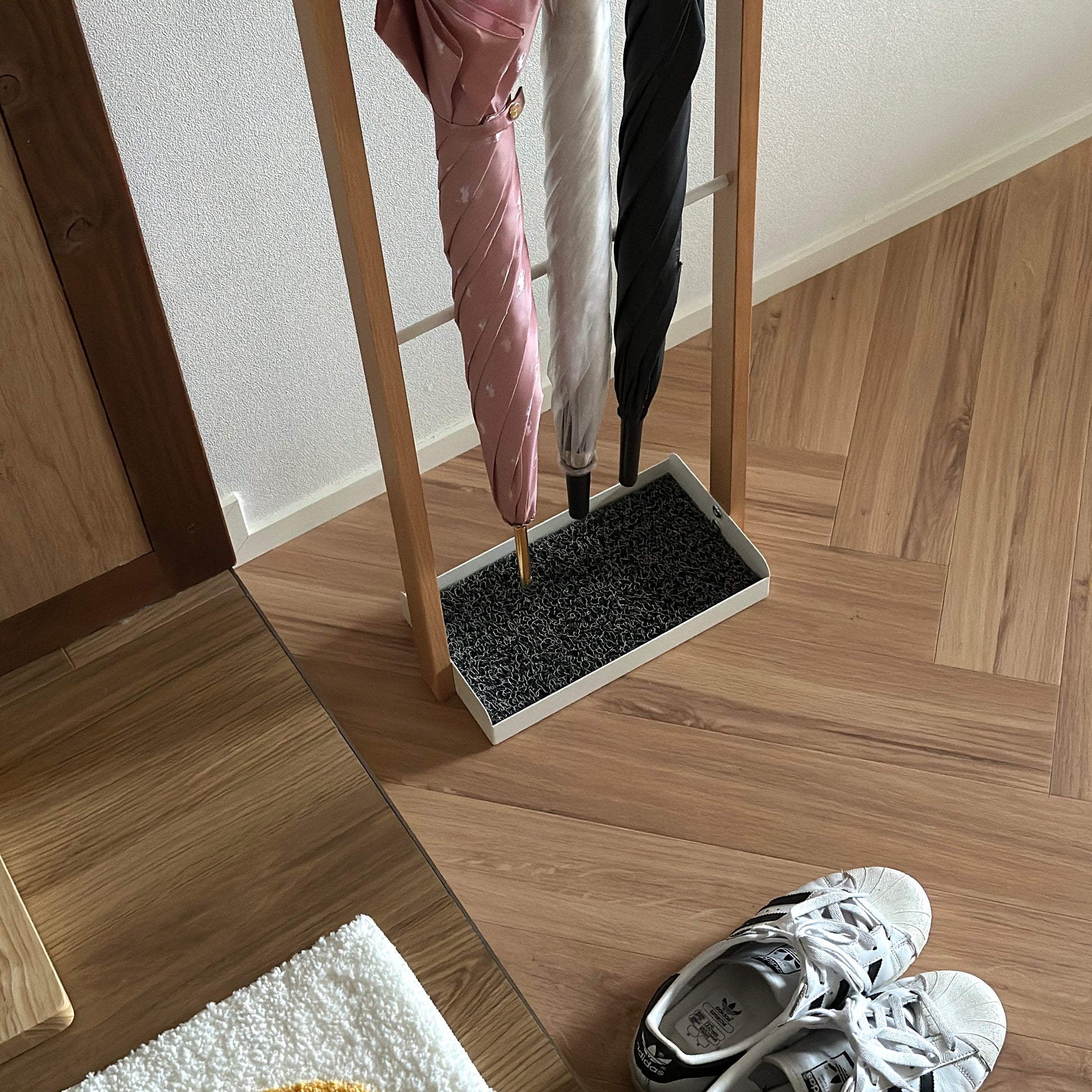 部屋の中に天然木とホワイトスチールの3段のアンブレララックが置かれており、ピンクの傘とビニール傘、黒の傘がかけられている。