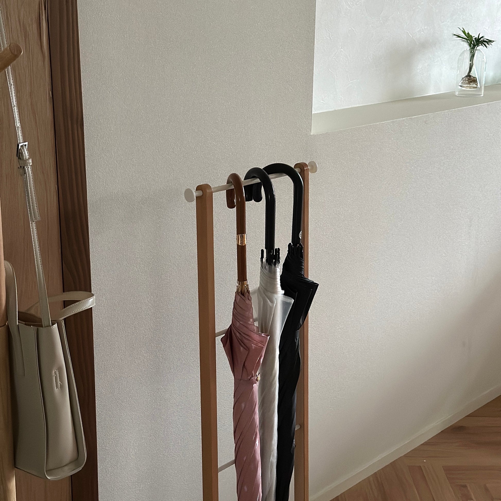 部屋の中に天然木とホワイトスチールの3段のアンブレララックが置かれており、ピンクの傘とビニール傘、黒の傘がかけられている。