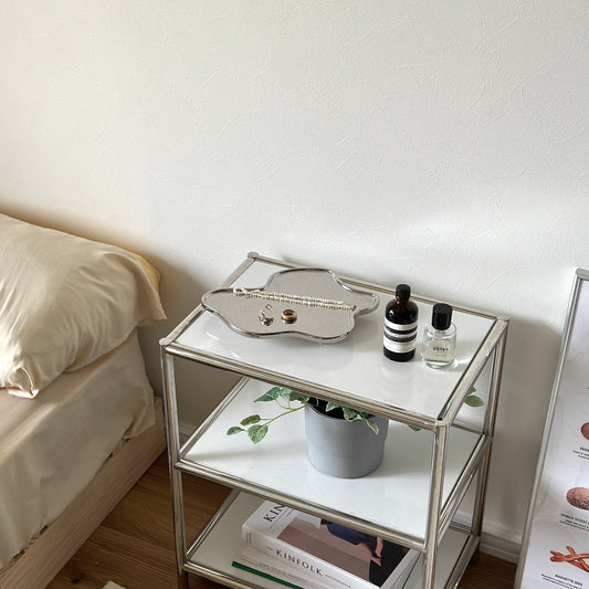 シルバーのフレームの白い棚の上に、縁がウェーブ状になっている脚付きのシルバープレートが置かれてある。