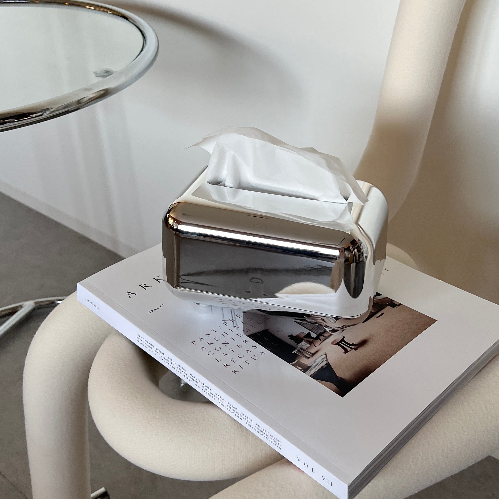 椅子の上に本の上に乗せられたツヤのあるメタルシルバーの脚付ティッシュケースが写っている写真